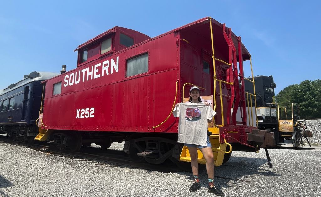 Image of woman holding t-shirt up in front of red caboose with "SOUTHERN X252" painted on it.
